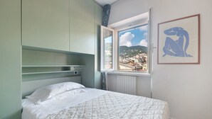 A bedroom with a bed, a window with a view of buildings and hills, and a framed artwork on the wall. - Apartment 'Appartamento Gabbiano' with Shared Terrace, Wi-Fi and Air Conditioning (Rapallo)