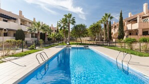 Una piscina con borde de baldosas, rodeada de palmeras y una valla, con edificios al fondo.