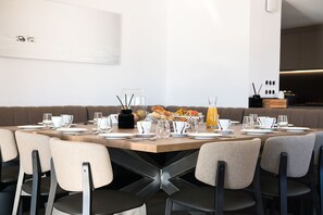 A modern dining area with a long table set for breakfast, including plates, glasses, and a fruit basket.
