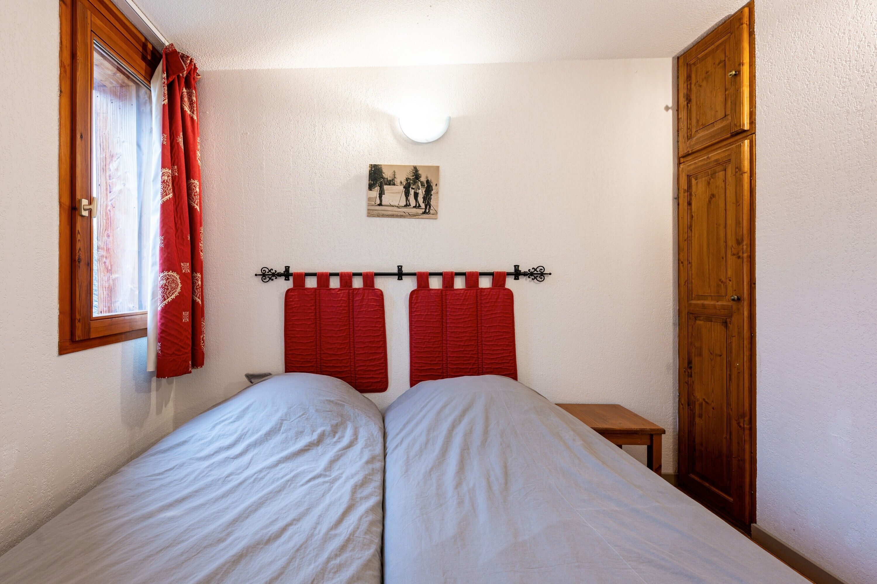 A bedroom with two beds, a wooden door, a small table, a window with red curtains, and a wall-mounted light fixture.