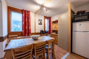 Una mesa de madera con sillas, una zona de cocina con un refrigerador y un ventanal con cortinas.