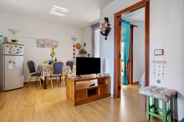 Un salon avec un meuble TV en bois, une table à manger avec des chaises, un réfrigérateur et un petit tabouret.