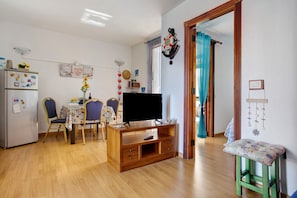 A living room with a wooden TV stand, a dining table with chairs, a refrigerator, and a small stool.