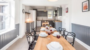 A dining area with a wooden table set for four, surrounded by black chairs. A kitchen is visible in the background with modern appliances and grey cabinetry. - The Red House, Leiston - A Smart Base for Coast and Work (Suffolk)