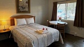 A hotel room with a bed, a small table, a chair, a window with curtains, and a wall-mounted picture. - Ashcroft Motel (Ashcroft)