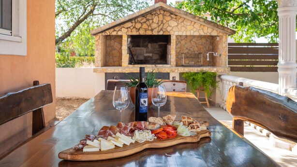 An outdoor dining area with a stone barbecue, a bottle of wine, and a charcuterie board. - Awesome home in Kosute (Kosute)