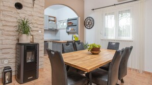 A modern kitchen with a stone wall, a dining area with a wooden table and leather chairs, and a large window with sheer curtains. - Awesome home in Kosute (Kosute)