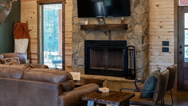 A cozy living room with a stone fireplace, a large flat-screen TV mounted above it, and a leather sofa. - Cozy 4 bedroom Lodge close to I-30 and local wedding venues (Simms)
