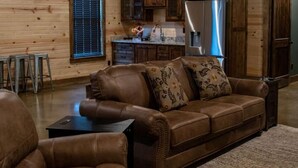 A cozy living room with a brown leather sofa, a coffee table, and metal stools. A kitchen with wooden cabinets and a refrigerator is visible in the background. - Cozy 4 bedroom Lodge close to I-30 and local wedding venues (Simms)