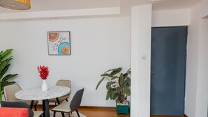 A dining area with a round table, chairs, a vase with red flowers, a painting on the wall, and a large potted plant.