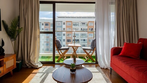 A modern living room with a red sofa, a wooden coffee table, and two chairs. A large window offers a view of a residential building.