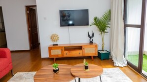 A modern living room with a flat-screen TV mounted on the wall, a wooden entertainment unit, two round coffee tables, a red sofa, and a large window with a view of a green lawn.