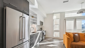A modern kitchen with stainless steel appliances, a microwave, and a built-in oven. There is a window with a view of the city, and a sofa with cushions in the living area. - Urban Elegance | 1BR LoHi Loft | City Views | Dog-Friendly (Denver)
