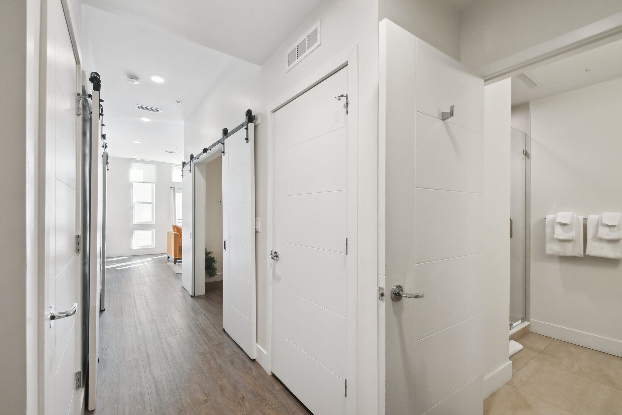 Un pasillo con puertas blancas, suelo de madera y vista a otra habitación con ventana y planta.