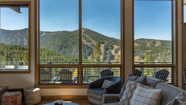 A cozy living room with large windows offering a mountain view, a comfortable sofa, and two Adirondack chairs.