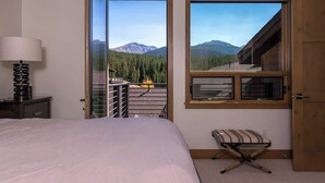 A bedroom with a bed, a nightstand, a lamp, a window with a mountain view, and a wooden door.