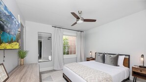 A bedroom with a bed, a ceiling fan, a window with curtains, a bedside table with a lamp, and a wall-mounted mirror.
