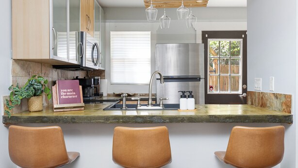 A modern kitchen with a granite countertop, stainless steel appliances, and a built-in oven. There are three brown chairs under the window.
