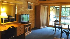 Una habitación con pared de ladrillo, un televisor sobre un soporte de madera, un escritorio con lámpara, una mesa de comedor con sillas y un ventanal con vistas a la vegetación.