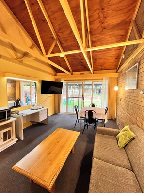 A room with a wooden ceiling, a TV mounted on the wall, a dining table with chairs, a sofa, and a coffee table. - High Country Motor Inn (Bright)