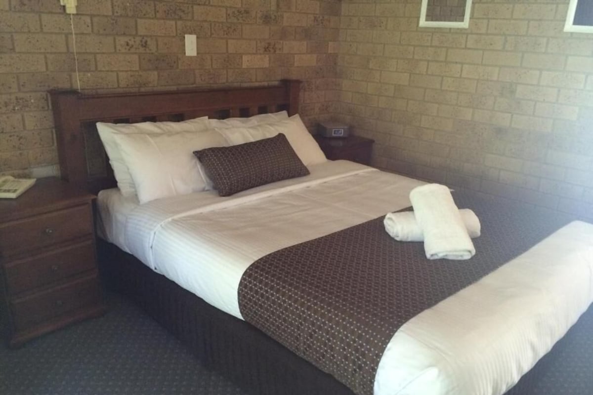 A neatly made bed with white linens and a brown blanket, a wooden headboard, and a brick wall.