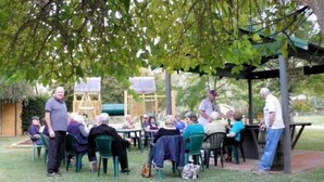 Reunión al aire libre con personas sentadas en mesas bajo un árbol, un área cubierta y un parque infantil al fondo.