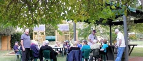 An outdoor gathering with people seated at tables under a tree, a covered area, and a playground in the background.