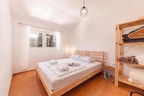 A bedroom with a wooden bed, a window with curtains, a hanging light fixture, and a wooden shelf with towels.