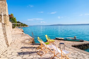 A stone-paved terrace with lounge chairs and a boat, overlooking a calm sea and distant hills.