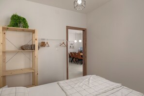 A bedroom with a bed, a wooden shelf with a plant, a mirror, and a dining area in the background.