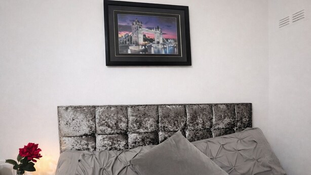 A bedroom with a stone-textured headboard, a framed picture of a bridge, and a vase with a red flower on a bedside table. - MCRShared House-Big Bedroom Sleeps 3 (Manchester)