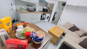 A compact kitchen with a sink, stove, and microwave. A dining table with food items and a basket is in the foreground. A sofa and a small wooden table are in the living area. - MCRShared House-Big Bedroom Sleeps 3 (Manchester)