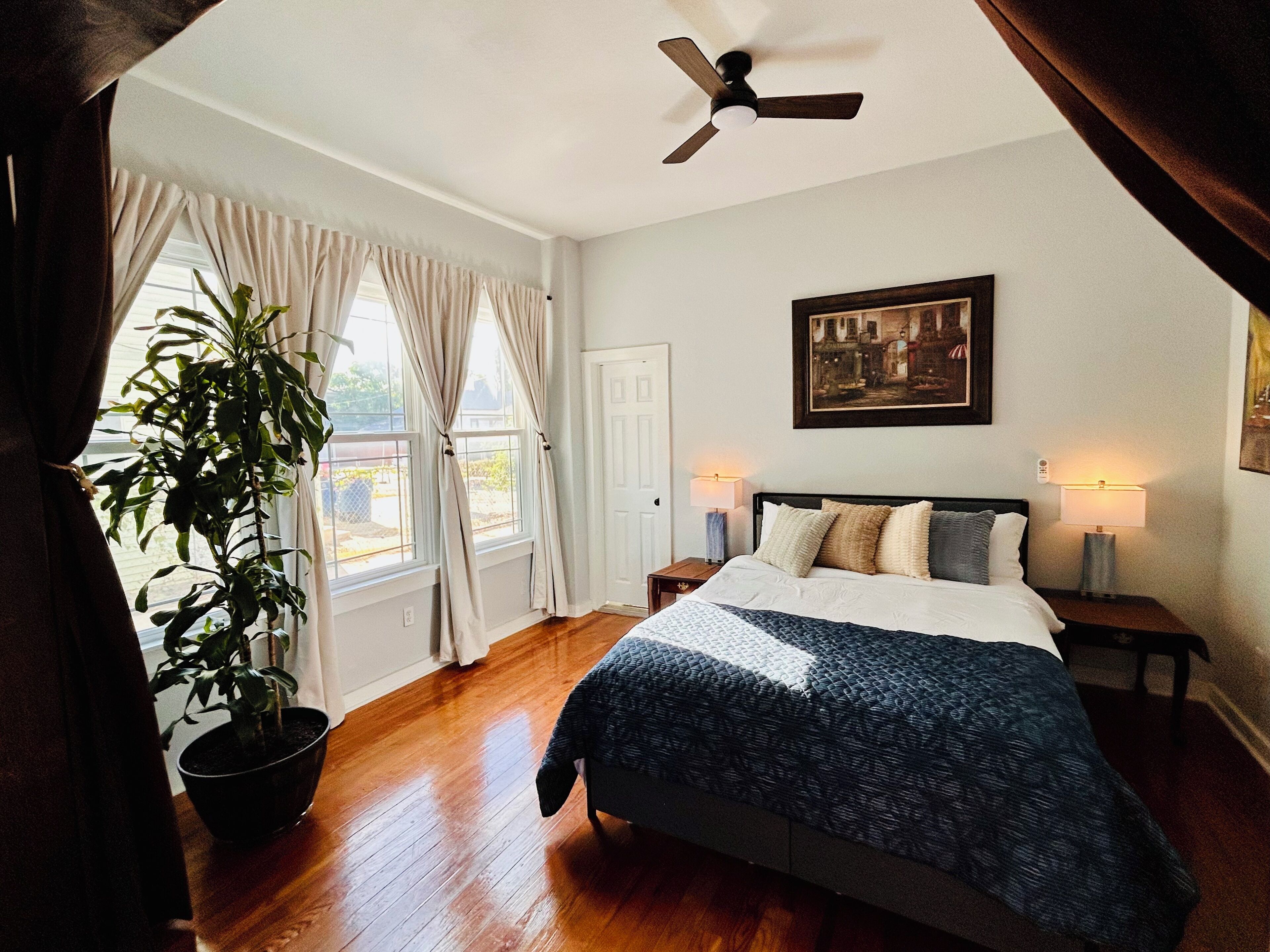 Une chambre à coucher avec un lit, un ventilateur de plafond, une table de chevet, une lampe, une plante en pot et une fenêtre avec des rideaux.
