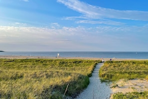 En strand med en græssti ned til vandet, et redder-tårn og folk i det fjerne.