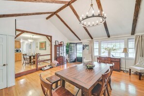 A spacious dining area with a wooden table and benches, a chandelier, and a view of the kitchen through a glass door. - 10 Mi to Crane Beach: Wooded Home Near Boston! (Topsfield)