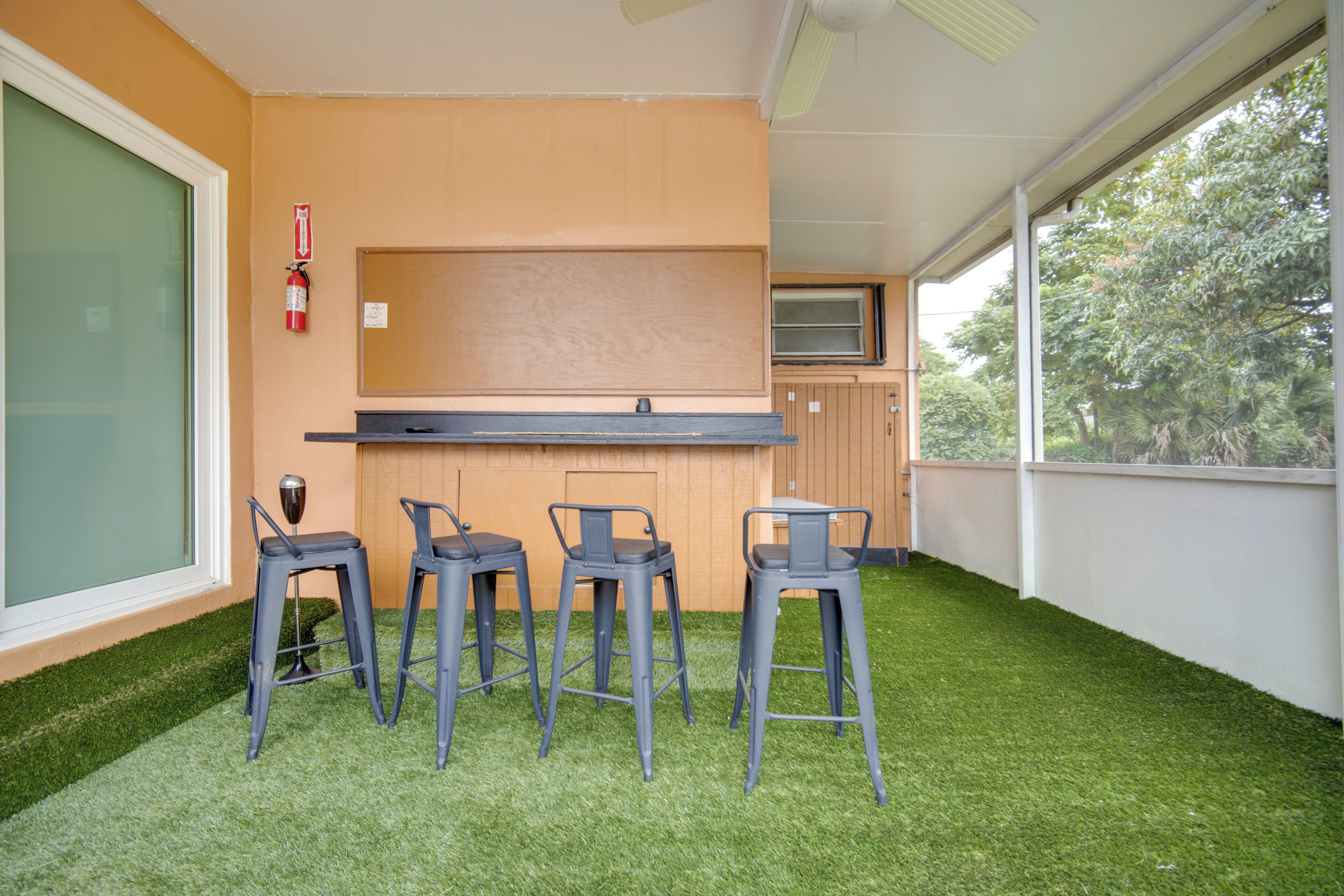 Un espace bar avec quatre tabourets et un comptoir, entouré d’un sol recouvert d’un gazon artificiel vert, et une fenêtre donnant sur des arbres.