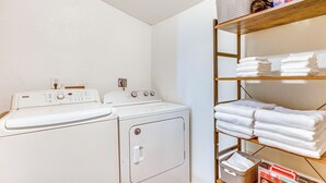 Un área de lavandería con lavadora y secadora de carga frontal, un estante con toallas dobladas y productos de limpieza.