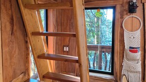 Interior de madera con escalera de madera, ventana con vista a árboles, un tapiz de macramé y una silla.