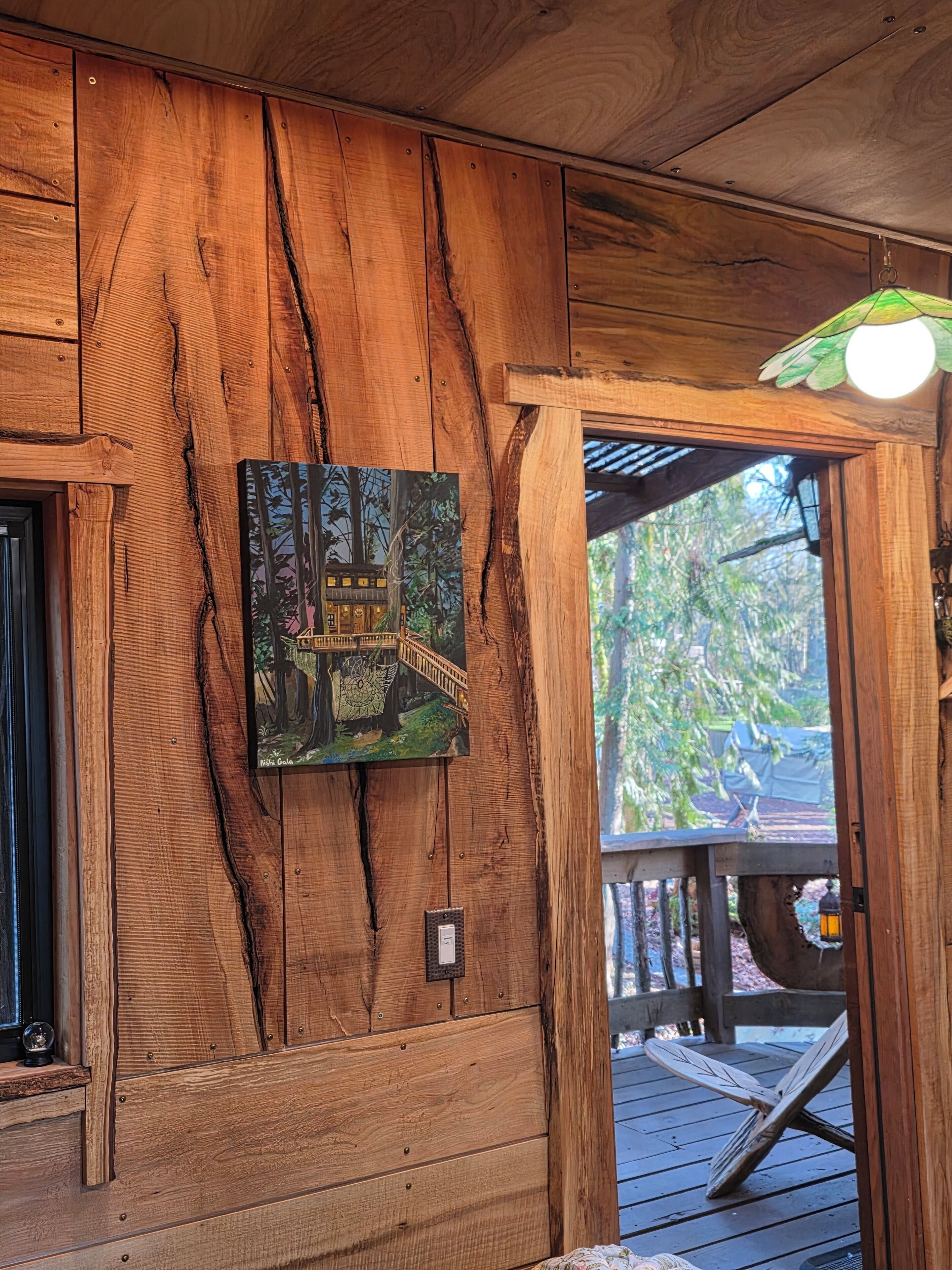 A wooden cabin interior with a framed painting of a treehouse on the wall, a wooden deck outside, and a lantern on the deck.