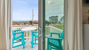 A balcony with turquoise chairs and a white table, overlooking a beach and a house.