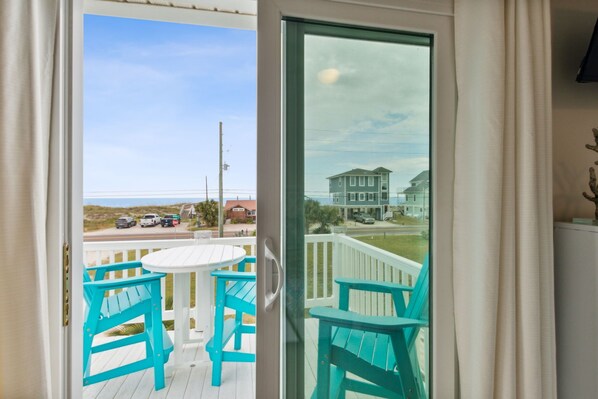 Un balcón con sillas turquesas y una mesa blanca, con vista a una playa y una casa.