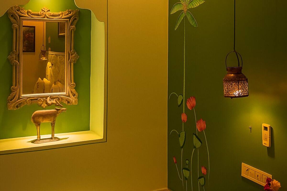 Une pièce avec un mur vert, un miroir doté d’un cadre orné, une petite statue, une lanterne décorative et un vase rempli de fleurs posé sur une table.