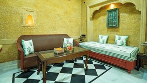 A room with a checkered floor, a brown sofa with decorative pillows, a wooden table, and a bed with a patterned bedspread. - Bhimkothi (Jaisalmer)