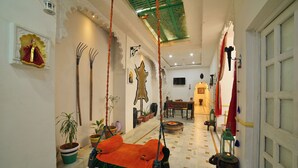 A corridor with a hanging swing, potted plants, a small table with a chair, and a door with a window. - Bhimkothi (Jaisalmer)