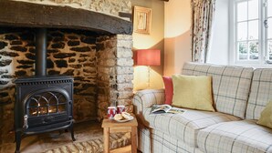A cozy room with a stone fireplace, a sofa with plaid cushions, and a window with curtains. - Strand Cottage - Burton Bradstock, Bridport (Burton Bradstock)