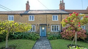 A stone house with a blue door, surrounded by well-maintained gardens and a paved pathway leading to the entrance. - Strand Cottage - Burton Bradstock, Bridport (Burton Bradstock)