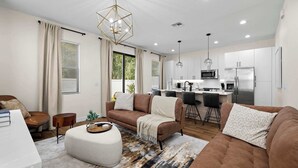 A modern living room with a brown sofa, a white ottoman, and a dining area with a kitchen in the background.
