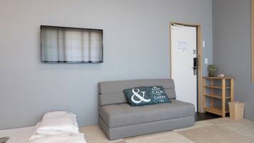 A room with a grey sofa, a bed with white bedding, a wall-mounted TV, a wooden door, and a small wooden shelf with a plant.
