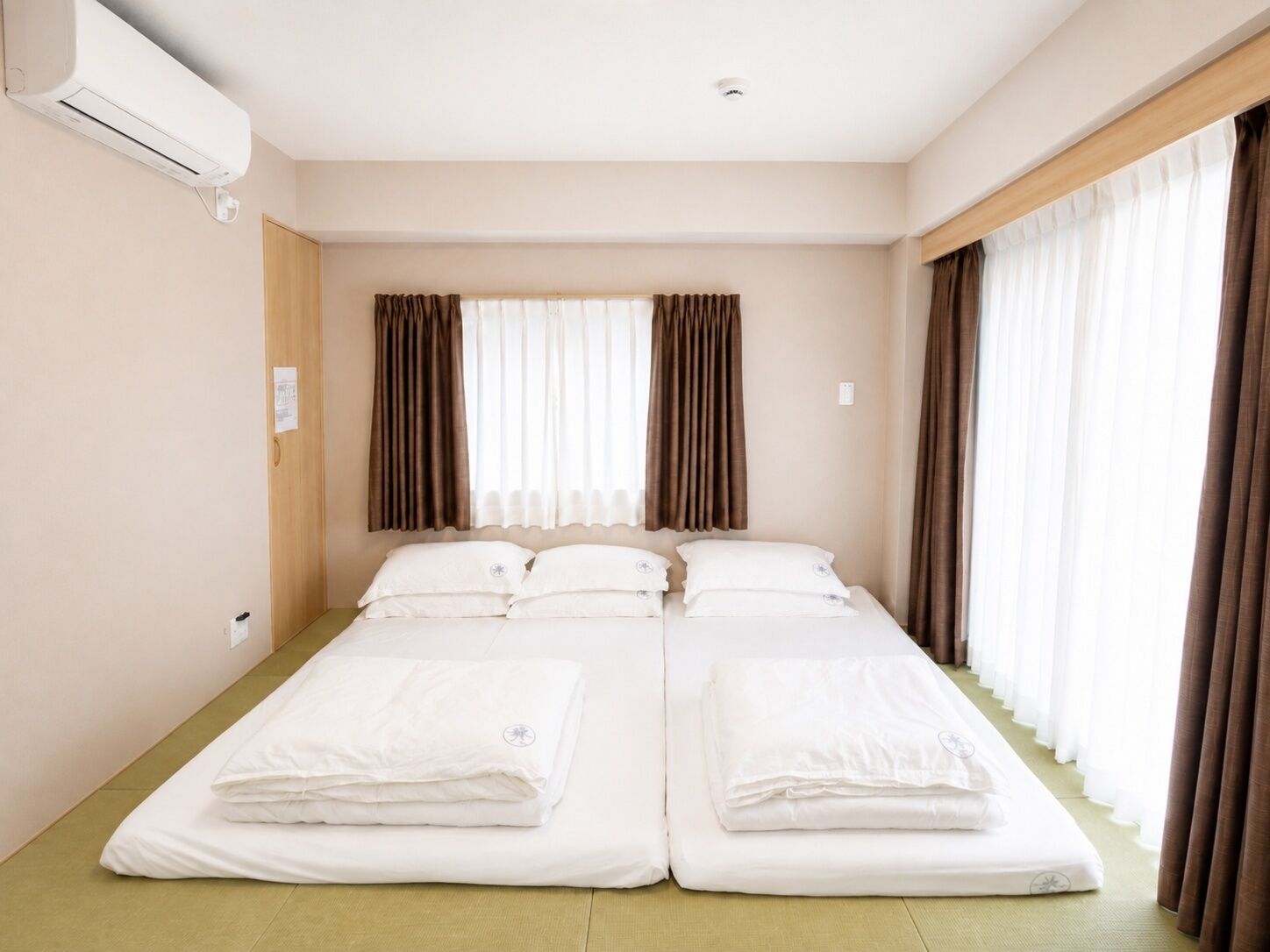 A hotel room with a double bed, a single bed, an air conditioning unit, and curtains on the windows.