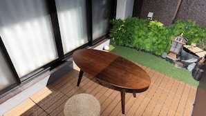 Een houten tafel met een rieten kruk op een betegelde vloer, nabij een raam met geëtste glas, een kleine tuin met groene planten en een decoratieve vogelhuis.